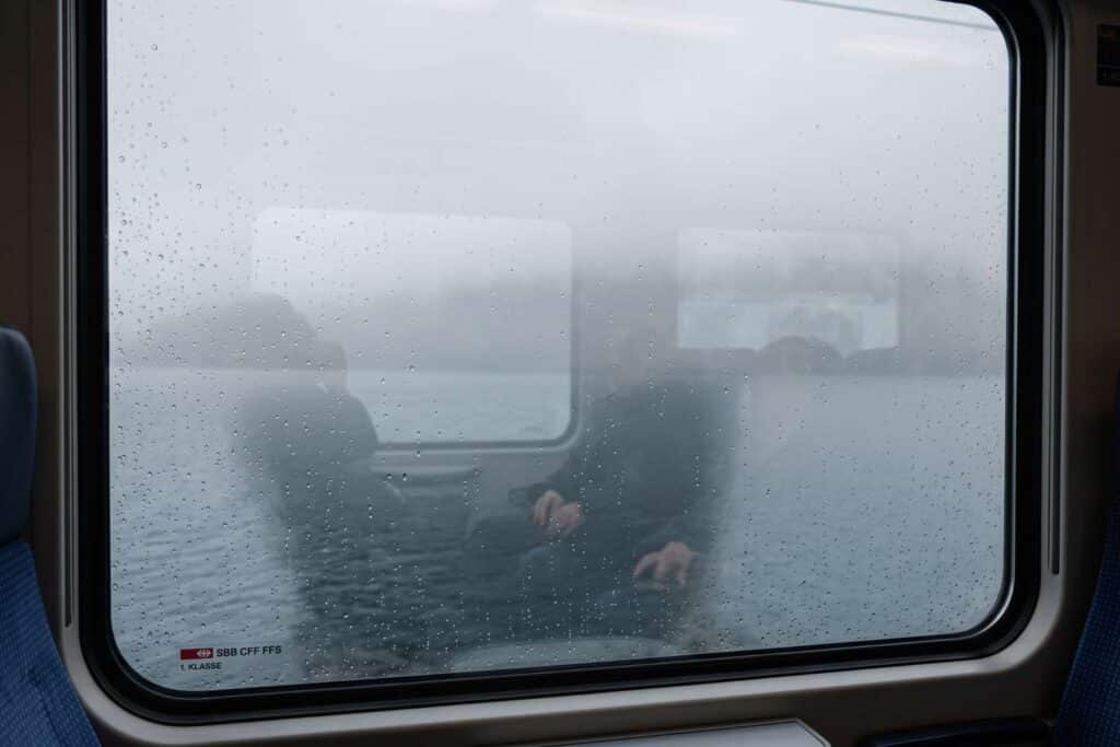Fenêtre de train suisse ruisselante de pluie ouvrant sur un lac noyé dans le brouillard d'hiver