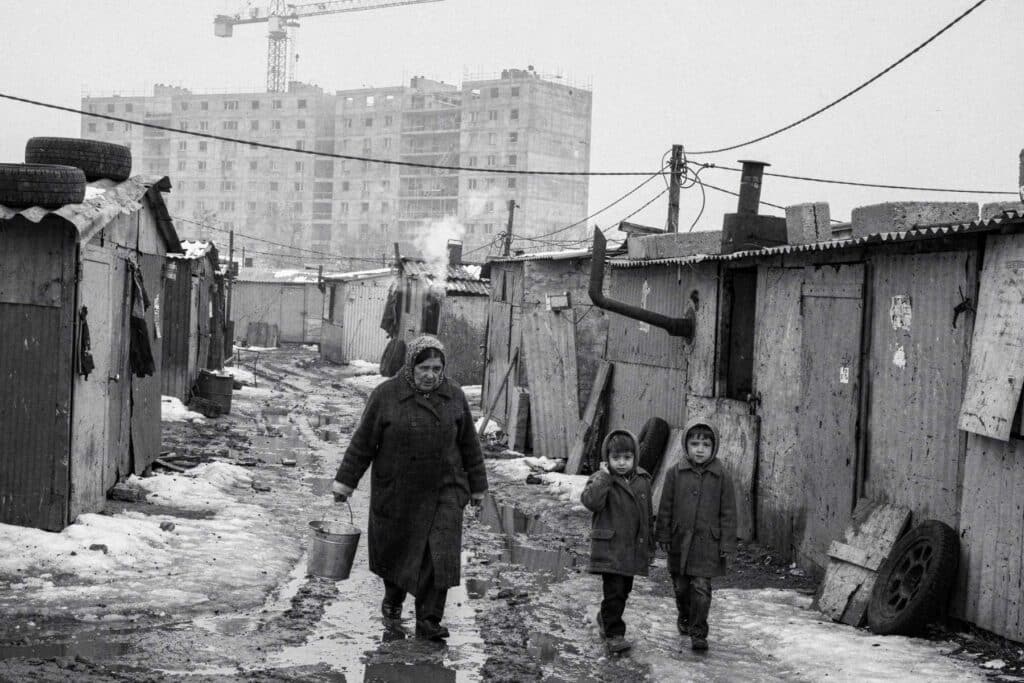 Bidonville de tôle sous la neige : une femme portant un seau d'eau et deux enfants, immeubles en construction à l'arrière-plan