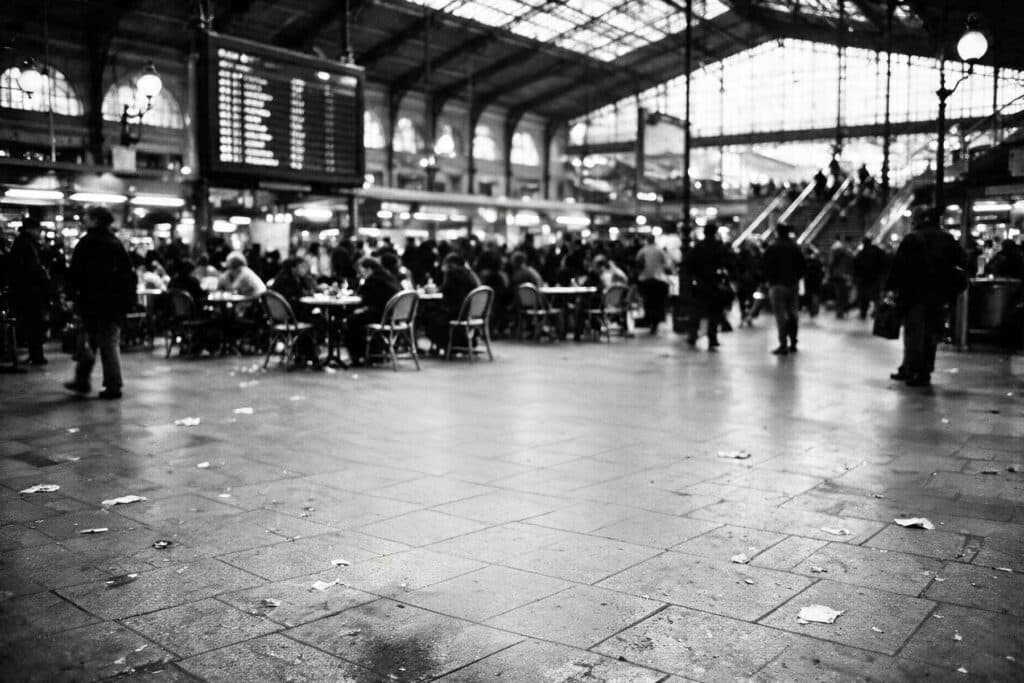 Hall d'une gare parisienne en noir et blanc, tables de café dressées au milieu d'une foule floue